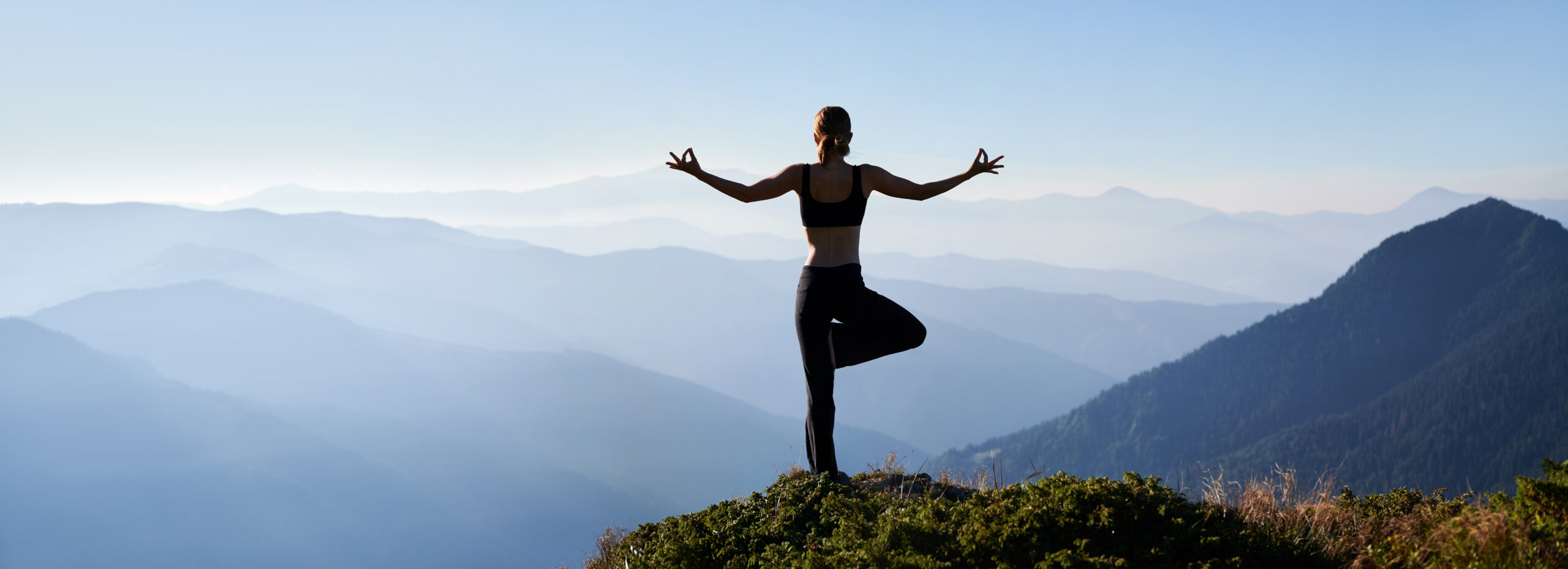 Frau macht Yoga auf einem Berg in der Natur, sie steht auf einem Bein und macht mit beiden Händen die klassische Yogapose
